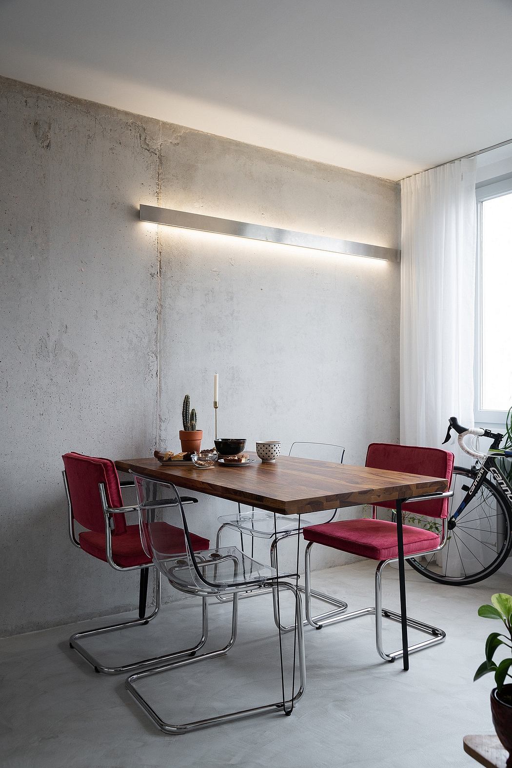 A minimalist dining area with a wooden table, red chairs, and a bicycle against a concrete wall.
