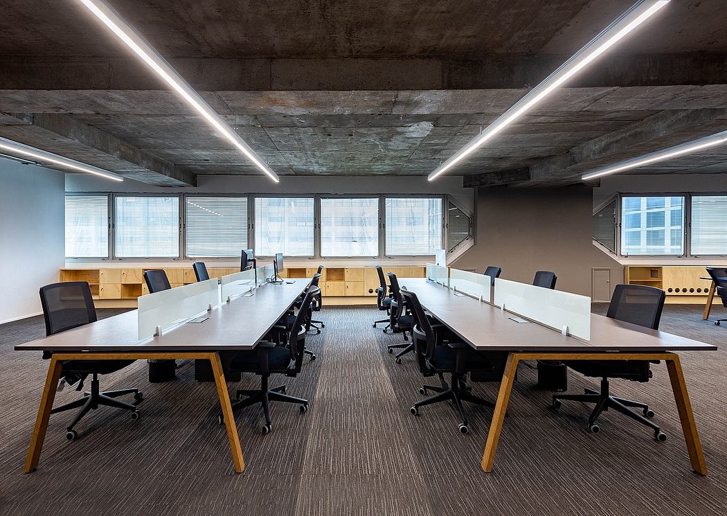 A spacious open-plan office with modern furniture, lighting, and exposed concrete ceiling.