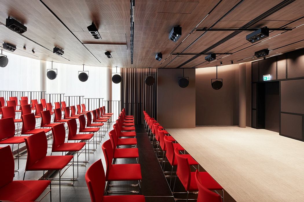 A modern auditorium with rows of red chairs, wooden ceiling, and theatrical lighting.