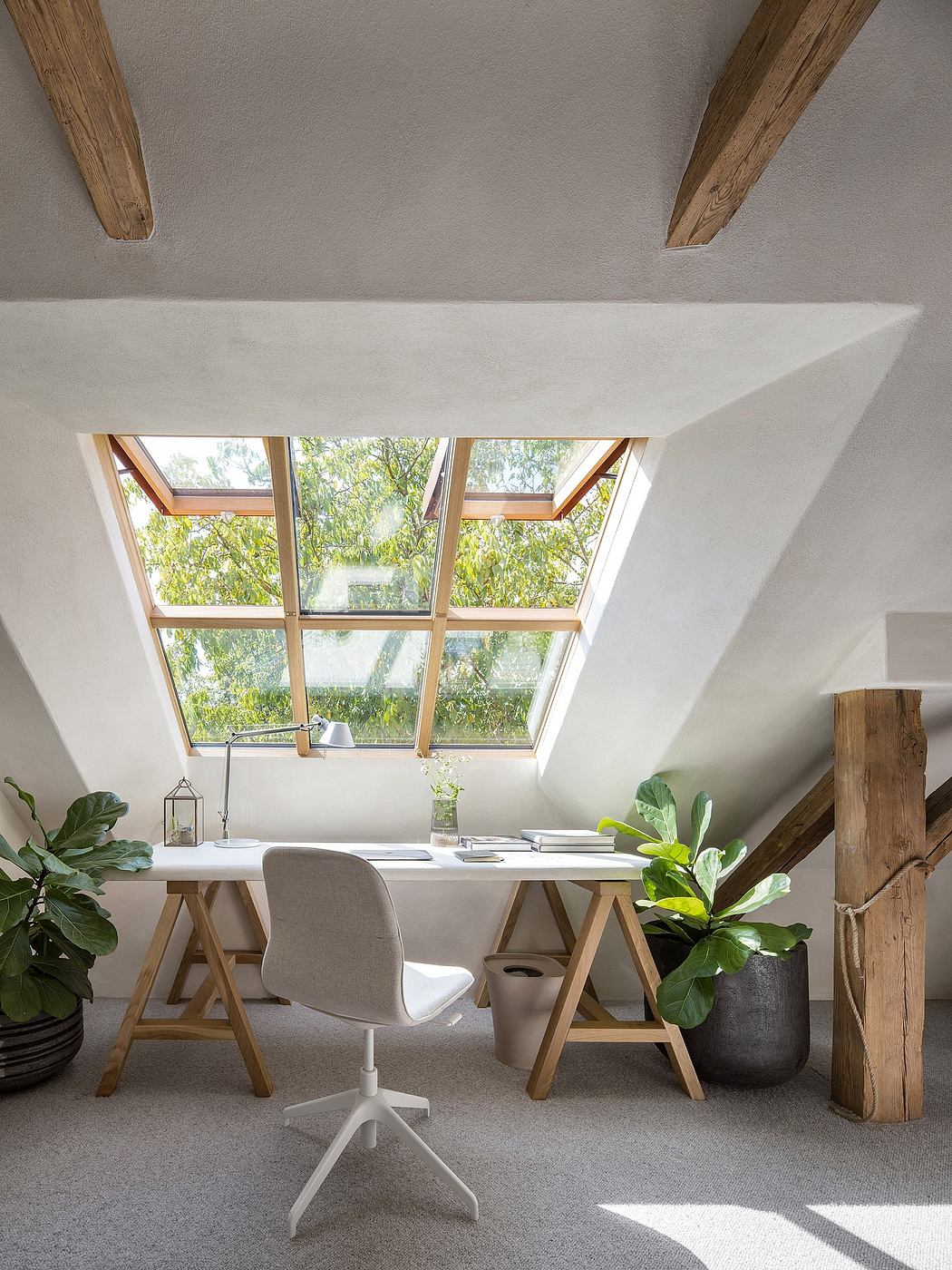 Cozy home office with skylight, wooden beams, and potted plants overlooking greenery.
