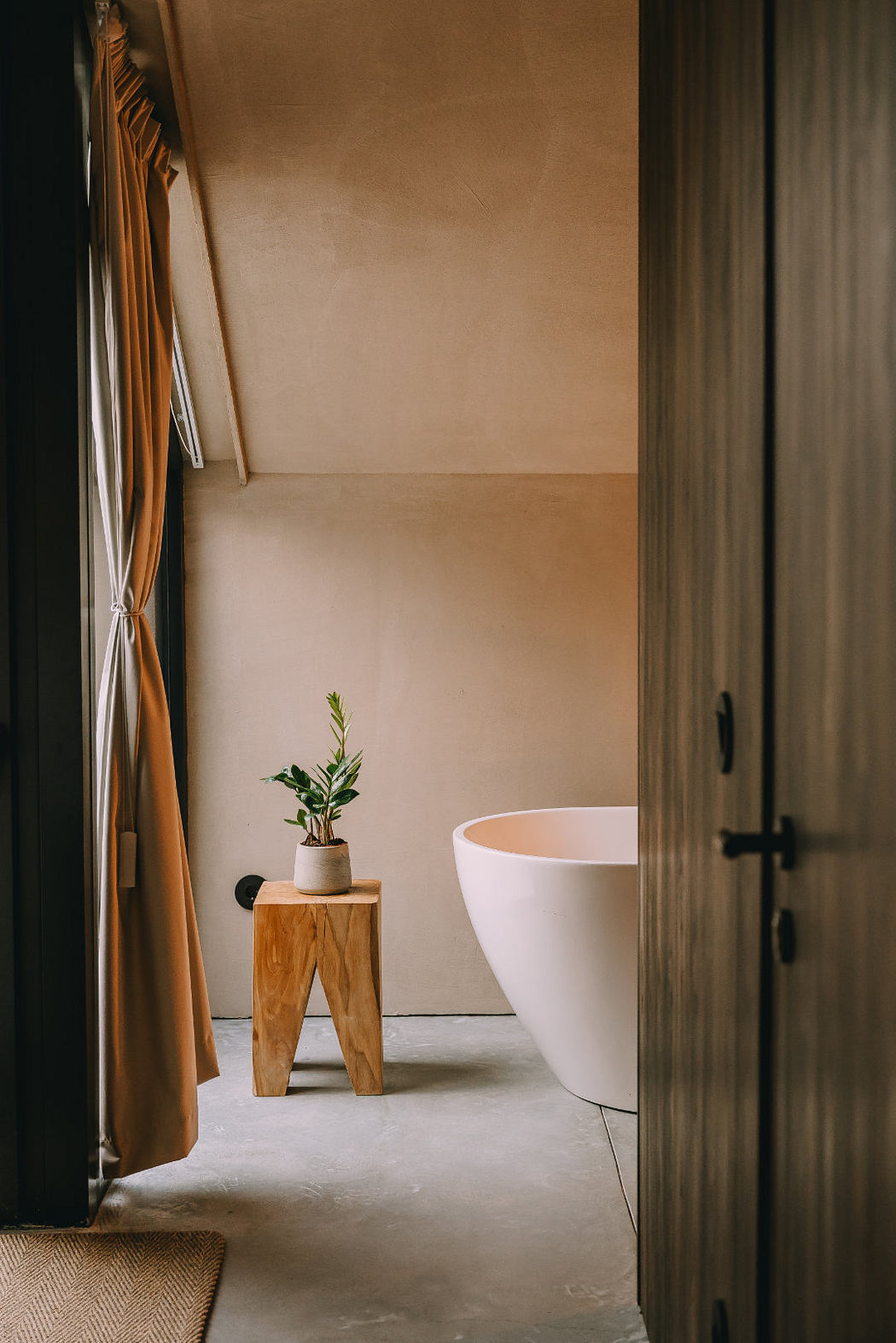 A cozy bathroom with a modern bathtub, wooden stool, and a potted plant, set in a warm, minimalist space.