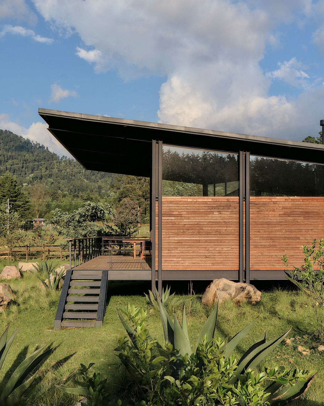 A modern, wooden cabin-like structure with a large overhanging roof and glass walls, surrounded by lush greenery.