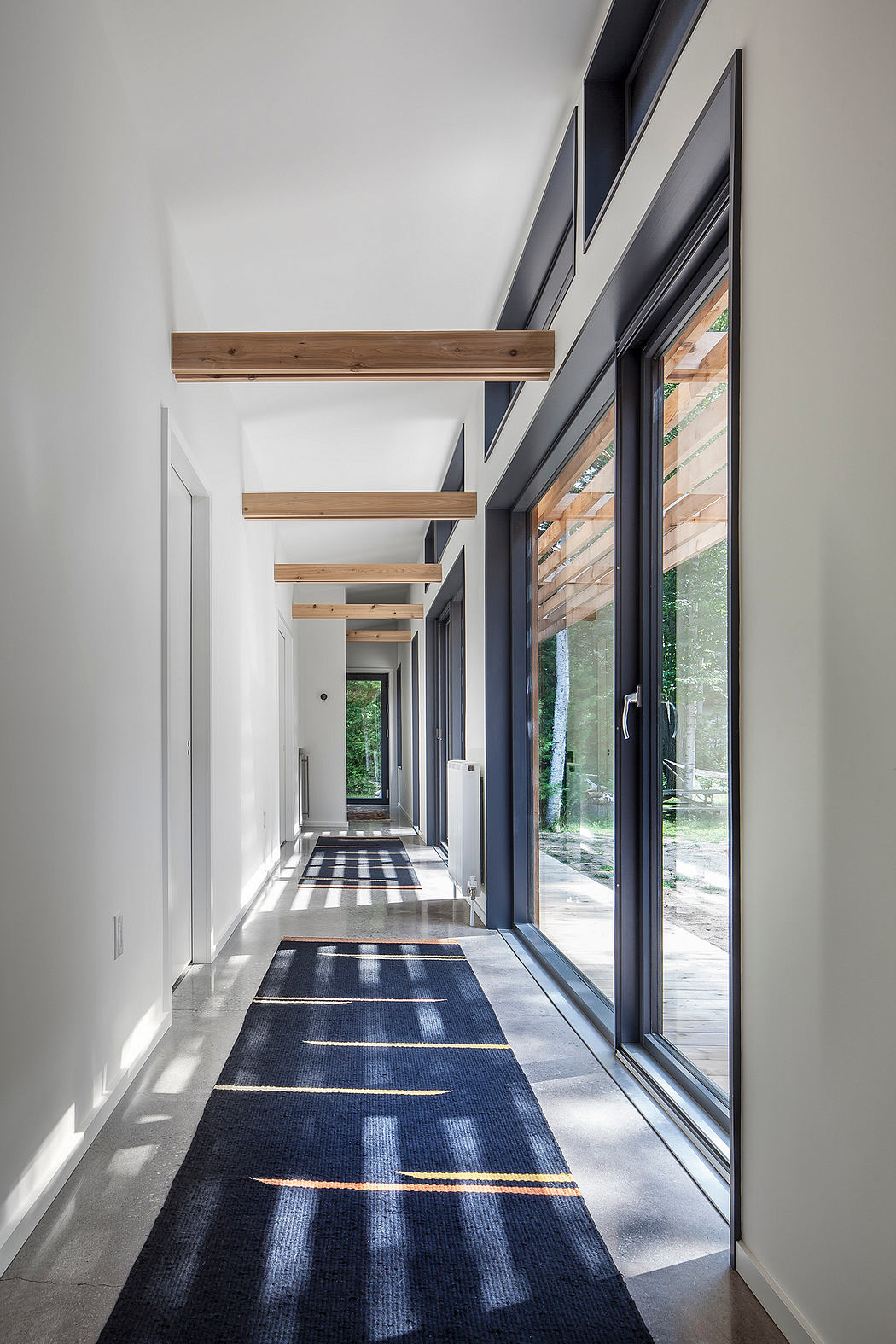 A modern, minimalist corridor with wooden beams, large windows, and a patterned floor rug.