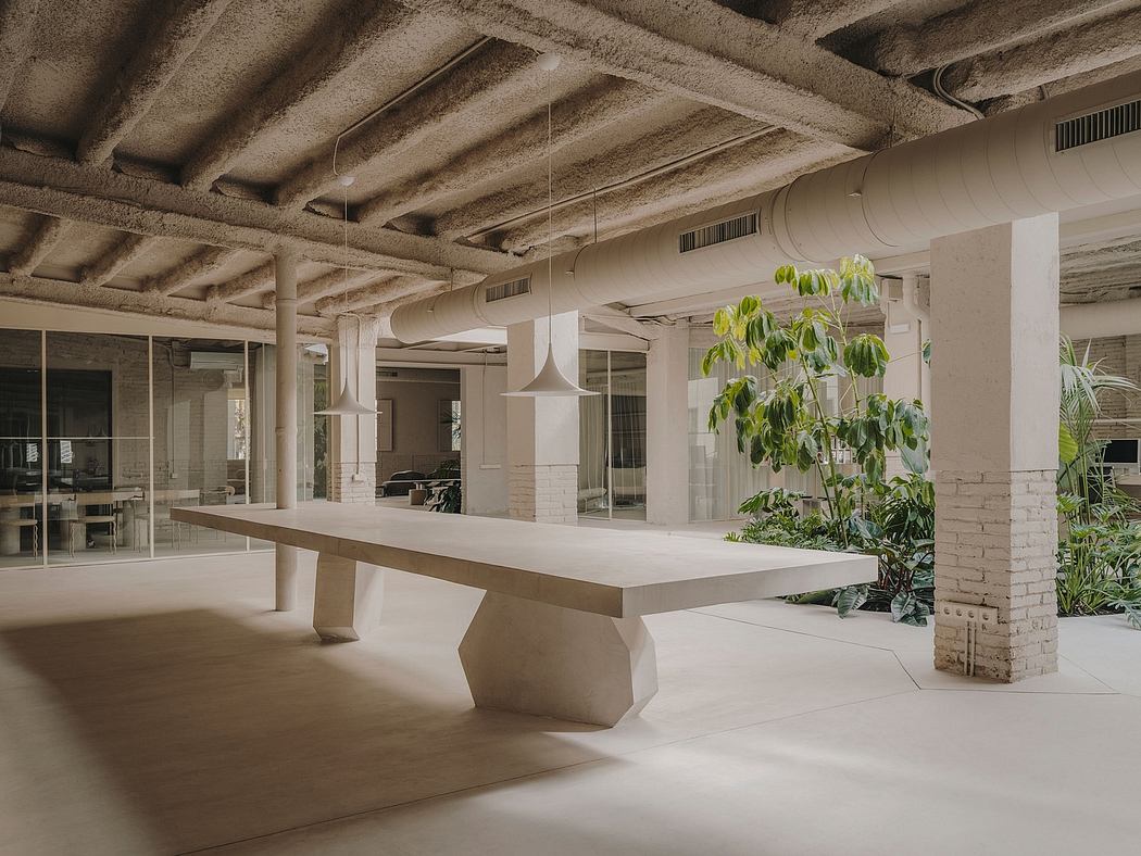 An open-concept industrial-style workspace with exposed beams, plants, and a long concrete table.