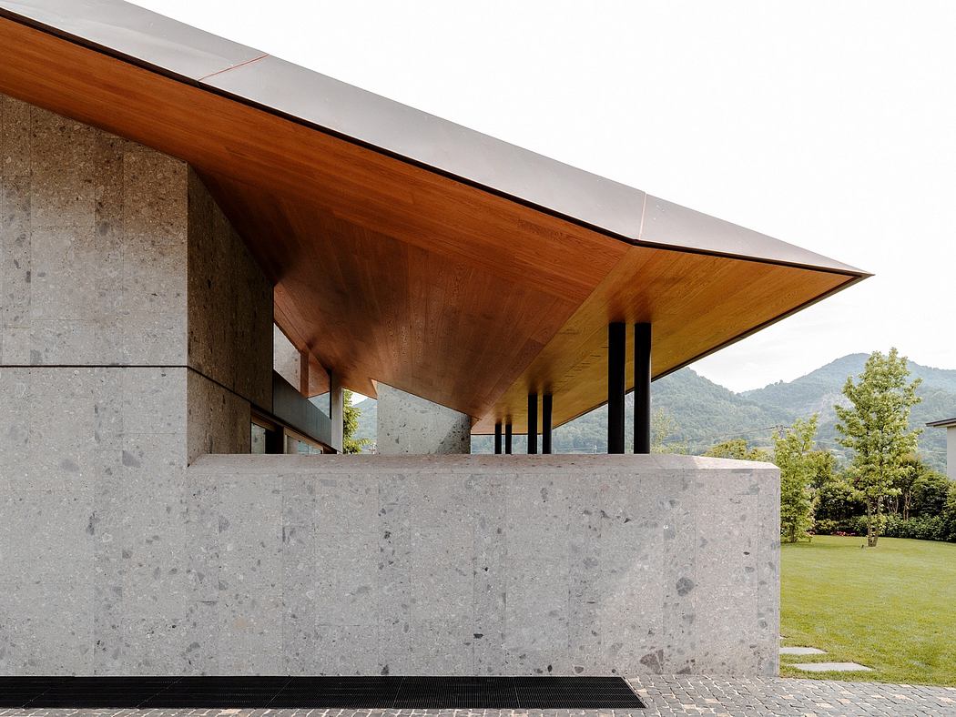A modern architectural design featuring a large wooden roof overhanging a concrete base, with a grassy landscape in the background.