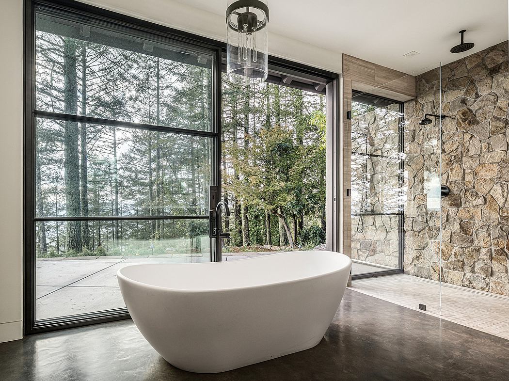 A modern bathroom with a freestanding bathtub, stone walls, and large windows overlooking a wooded landscape.