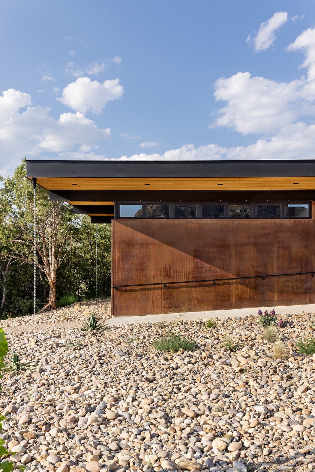 Rustic modern home features exposed wood beams, metal siding, and gravel landscape.
