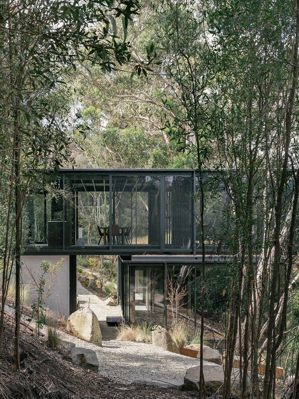 A modern glass and steel cabin nestled among lush greenery, with a path leading to its entrance.