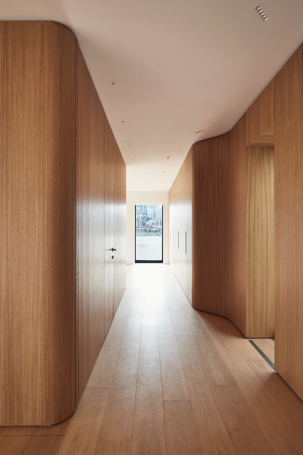 A well-lit, minimalist hallway with warm wood paneling and a glass door offering a view.