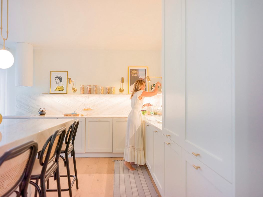 A cozy, sun-lit kitchen with white cabinetry, wooden floors, and an elegant woman examining the contents of the fridge.