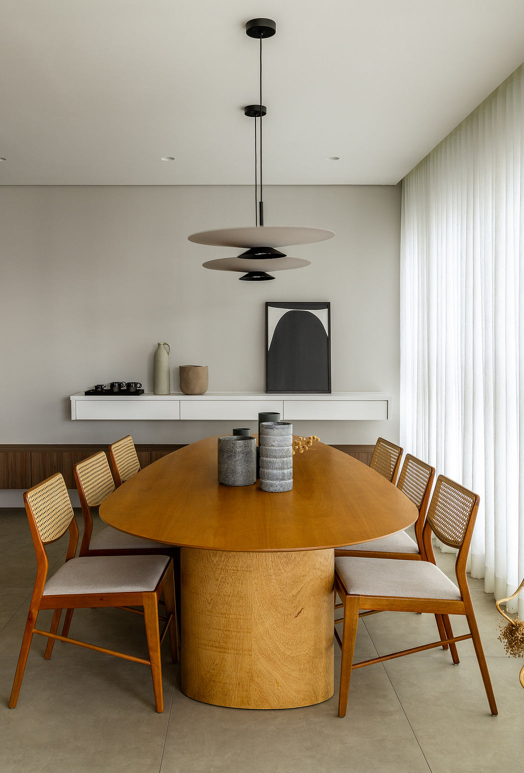 Modern dining area with a large round wooden table, caned chairs, and a minimalist hanging light fixture.