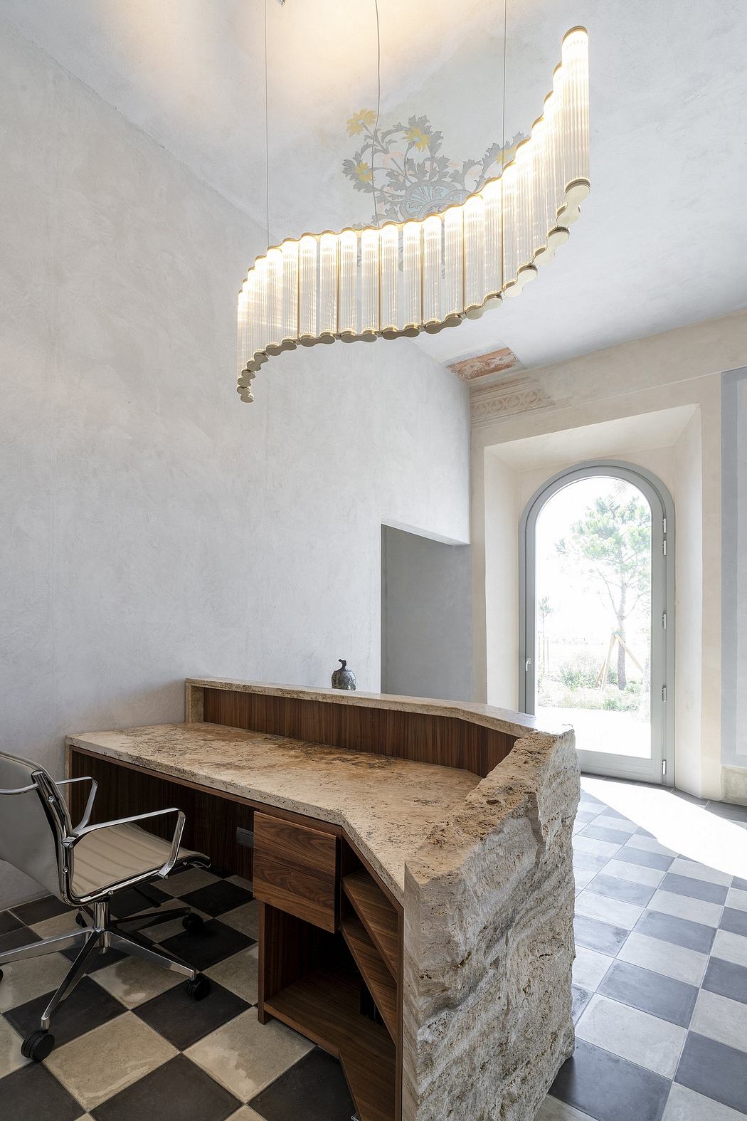 A modern office space with a large ornate light fixture, a wooden desk, and a window offering a scenic view.