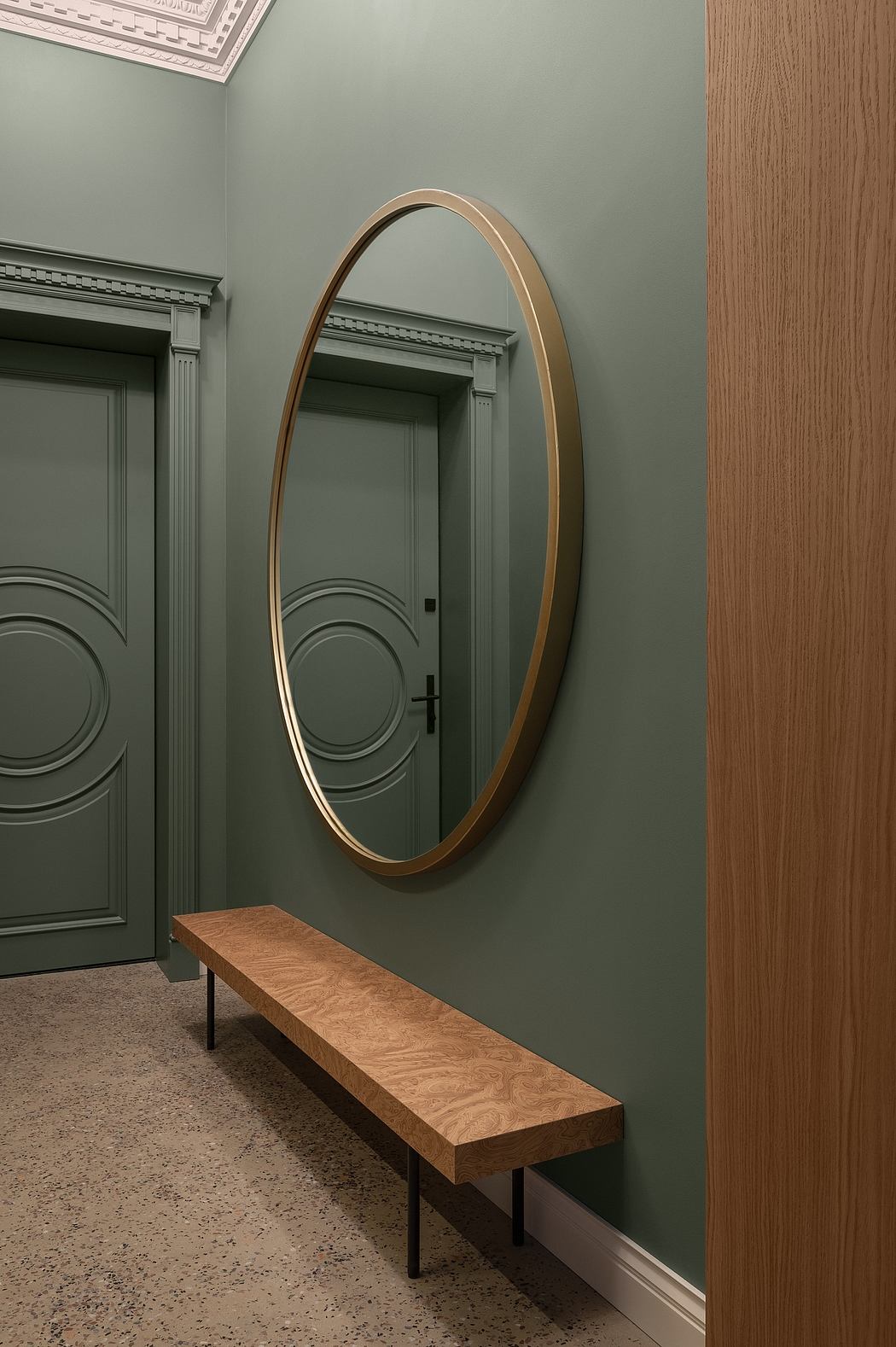 Elegant oval mirror, wooden bench, and green walls in a refined interior.