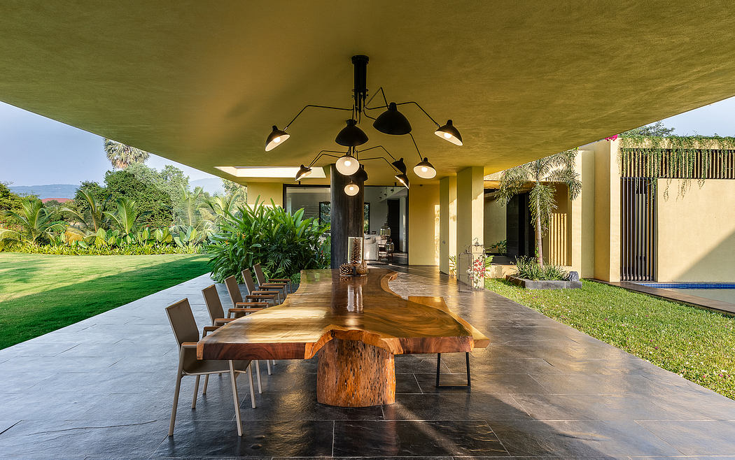 A modern, open-air dining area with a large wooden table, pendant lighting, and lush tropical foliage.