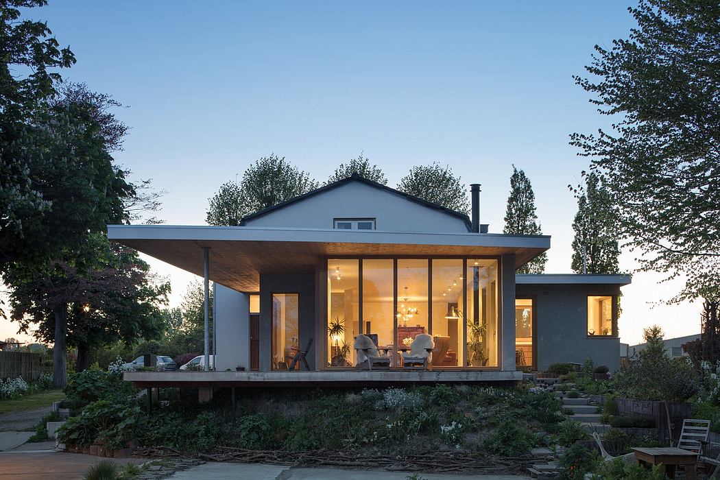 A modern, open-concept house with a large, wraparound deck set in a lush, wooded garden.