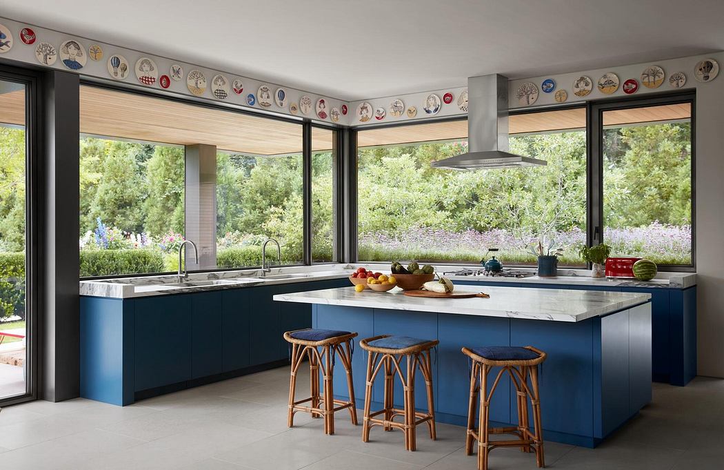 Bright, airy kitchen with large windows overlooking lush vegetation. Stylish blue cabinetry, wooden stools, and decorative plates on the walls.