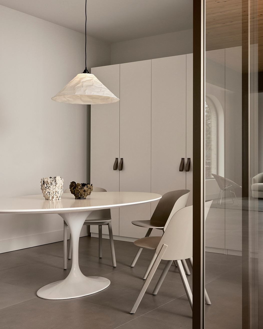 A modern, minimalist dining area with a white tulip table, wood chairs, and a fabric pendant light.