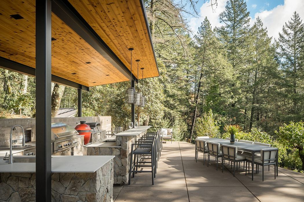 Outdoor kitchen and dining area with wooden ceiling, stone walls, and surrounding pine forest.