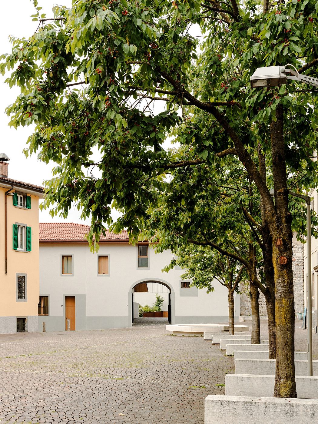 The image depicts a scenic square with a tree-lined courtyard, surrounded by buildings with arched entryways and residential facades.