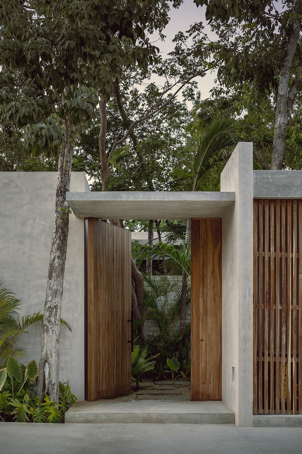 Minimalist concrete entryway with wooden doors and slatted screens, surrounded by lush greenery.