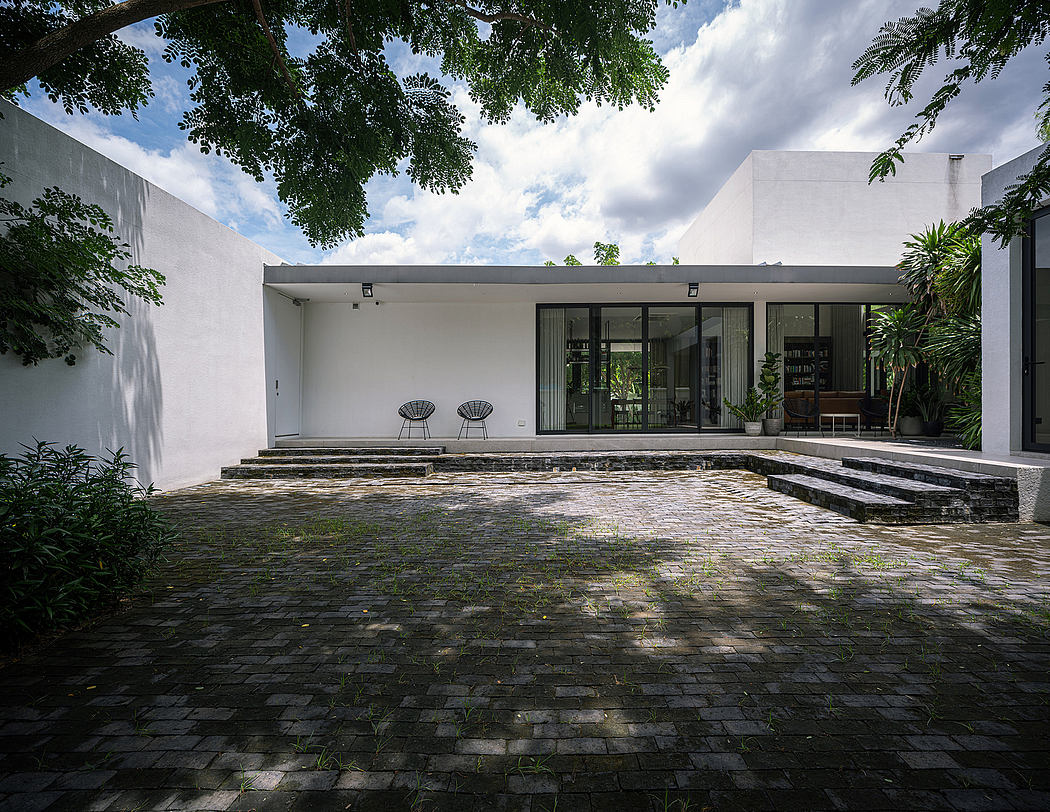 A modern, minimalist house with a paved courtyard, large windows, and lush vegetation.