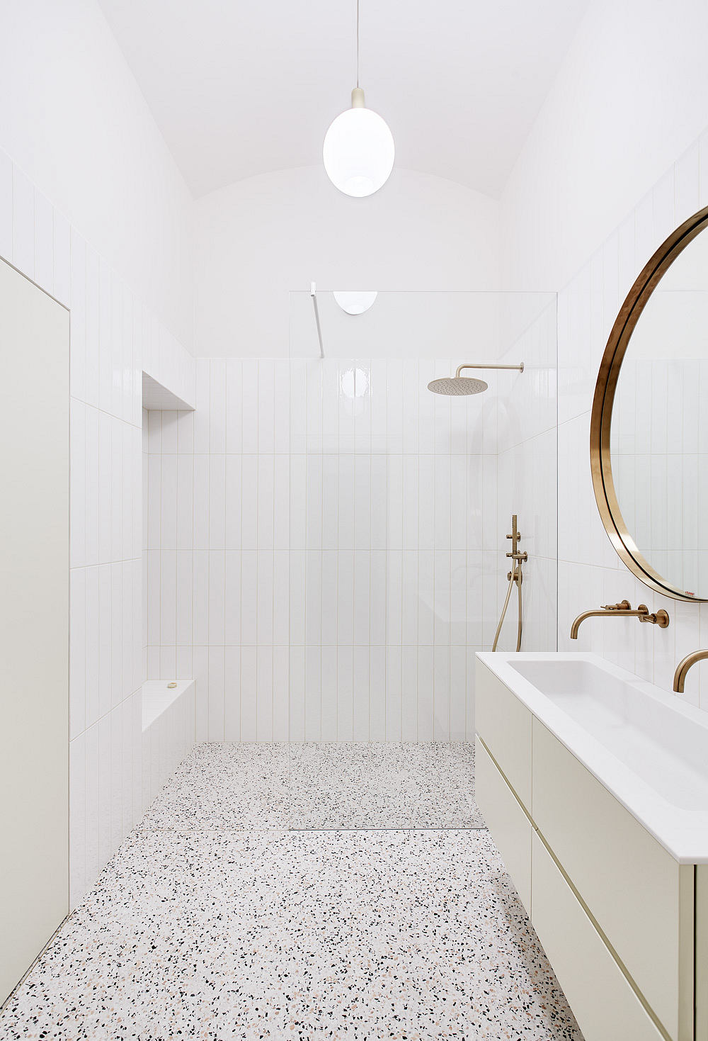 Minimalist bathroom with terrazzo flooring, brass fixtures, and circular mirror.