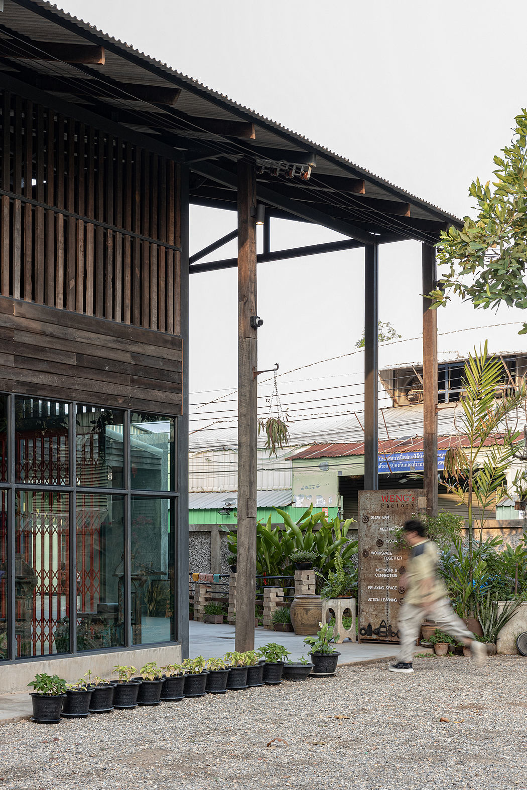 A rustic wooden structure with a covered patio, featuring potted plants and a blurred figure in the foreground.