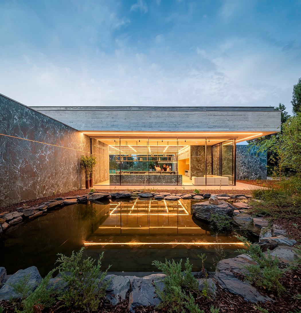 A modern, glass-walled structure with a stone wall and a pond reflecting its warm, inviting interior.