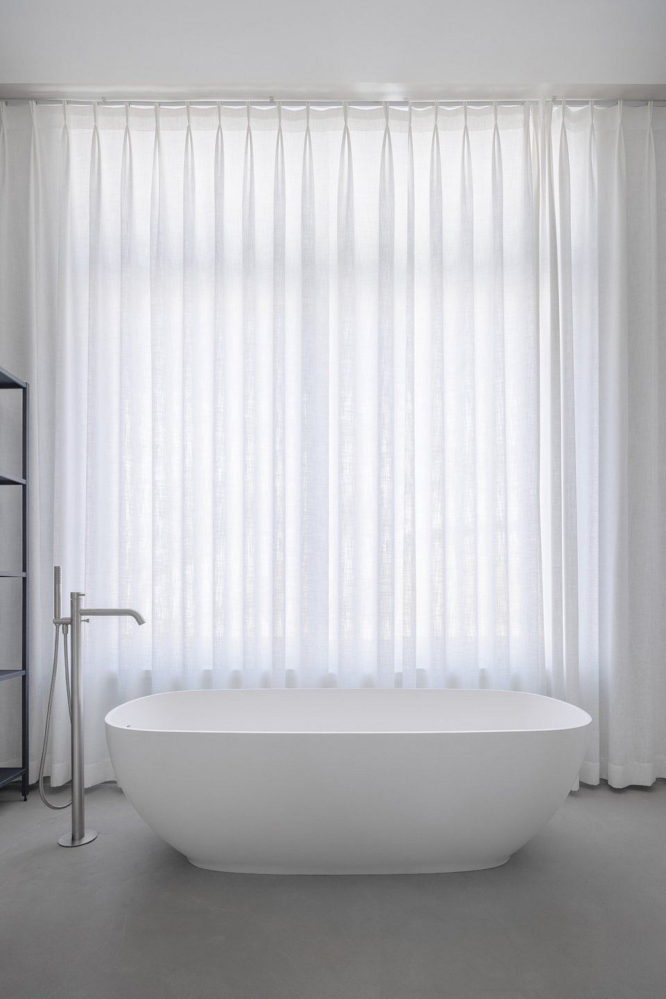 A minimalist bathroom with a freestanding white tub, sheer curtains, and a chrome faucet.