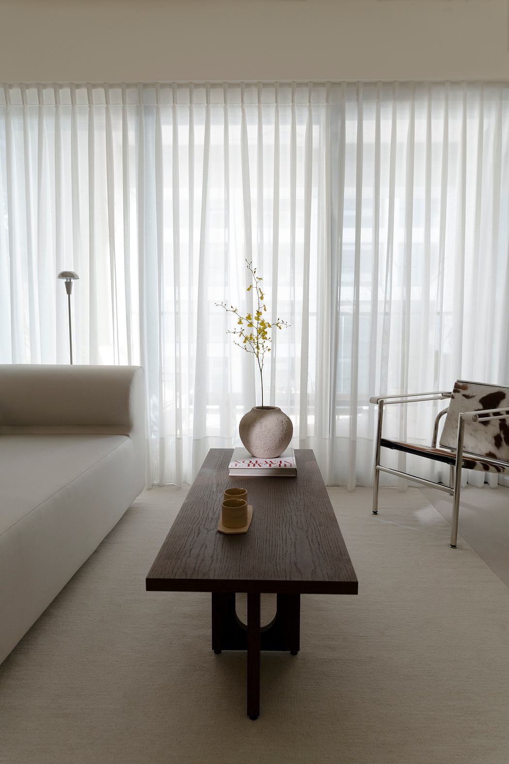 Minimalist living room with floor-to-ceiling curtains, wooden coffee table, and floral vase.