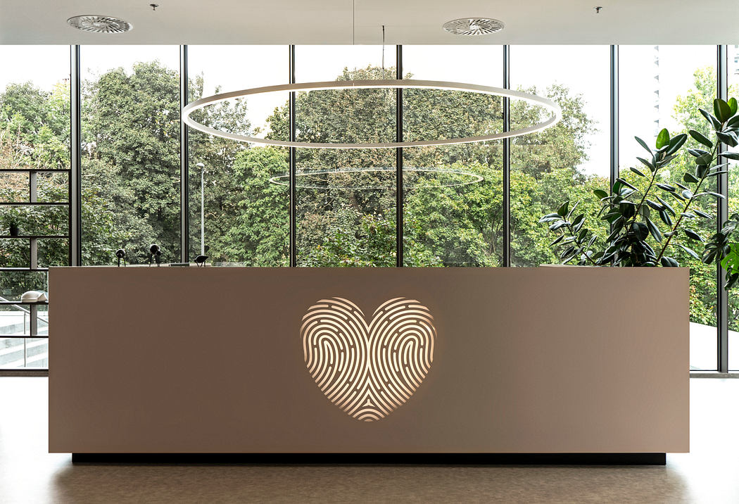 A modern lobby interior with a large, illuminated heart-shaped design on the reception desk, surrounded by floor-to-ceiling windows overlooking a lush, green landscape.