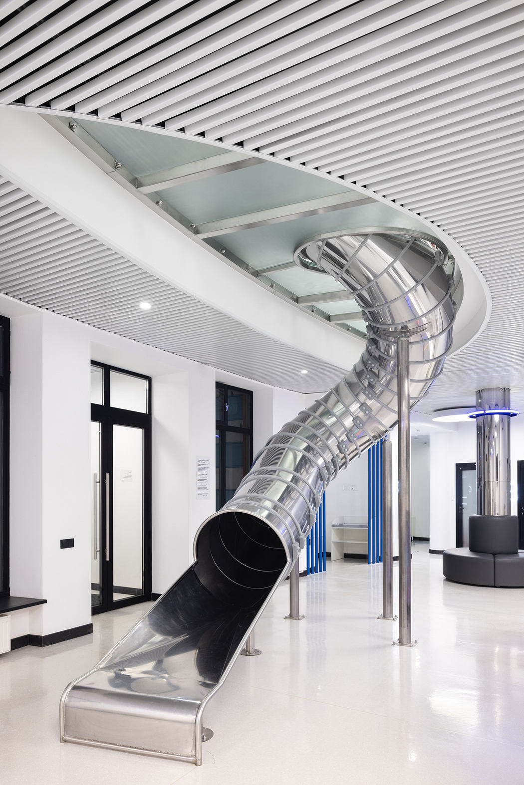 A sleek, modern indoor slide with a curved glass enclosure, surrounded by a striped ceiling.