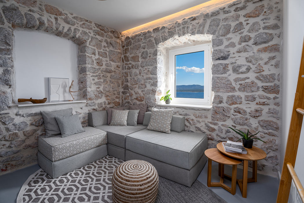 Cozy stone-walled living room with plush gray sectional, round woven pouf, and scenic window.
