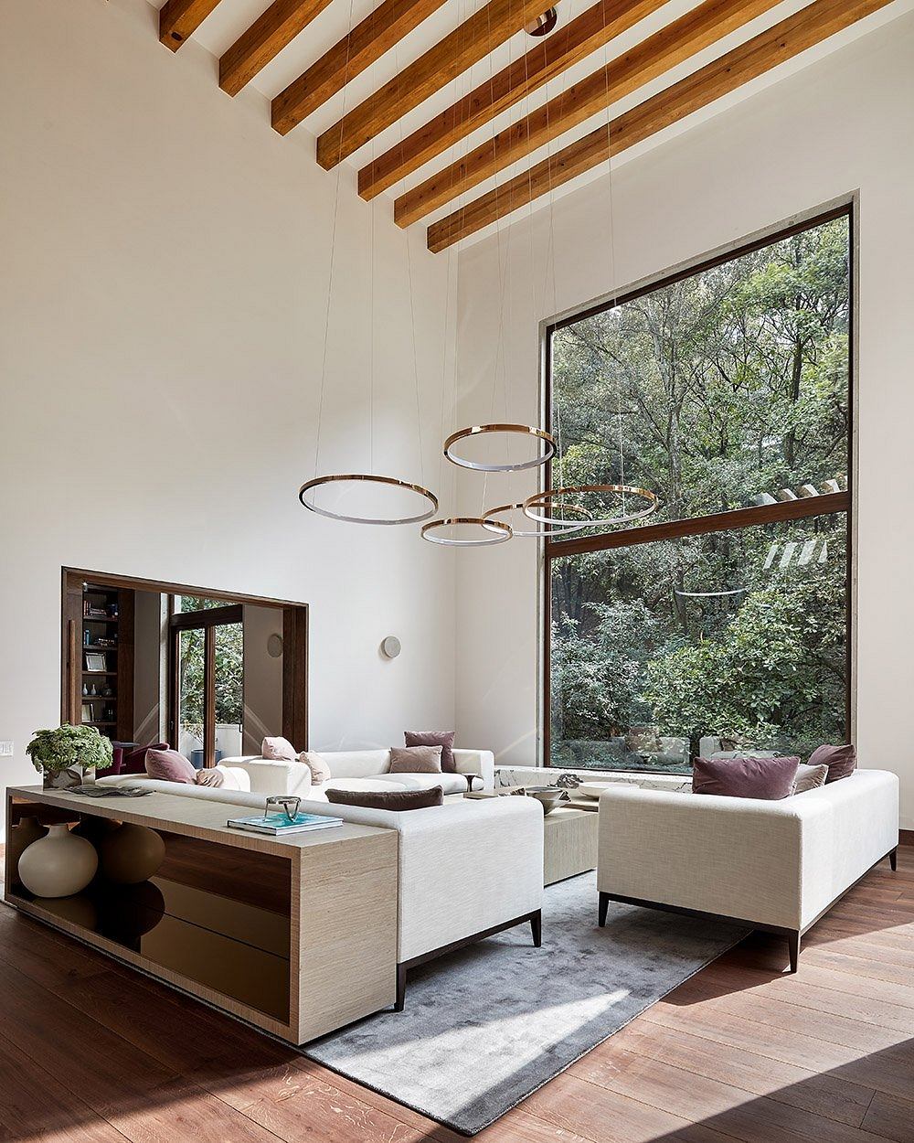 Spacious living room with wooden beams, circular pendant lights, and floor-to-ceiling windows overlooking greenery.