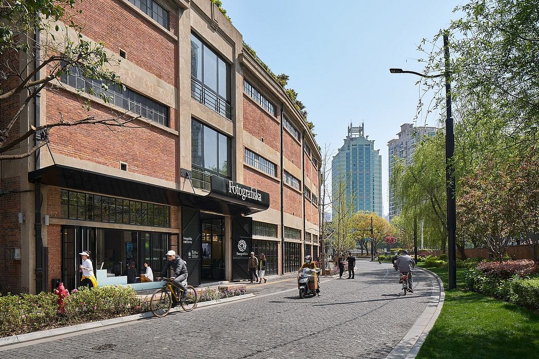 Brick building with large windows, trees along the sidewalk, and pedestrians on a sunny day.
