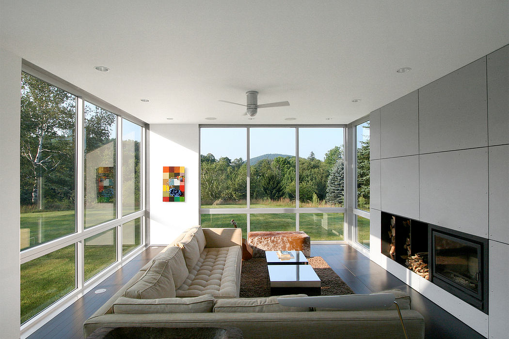 Large windows overlook a lush green lawn and trees. Modern, minimalist interior with fireplace.