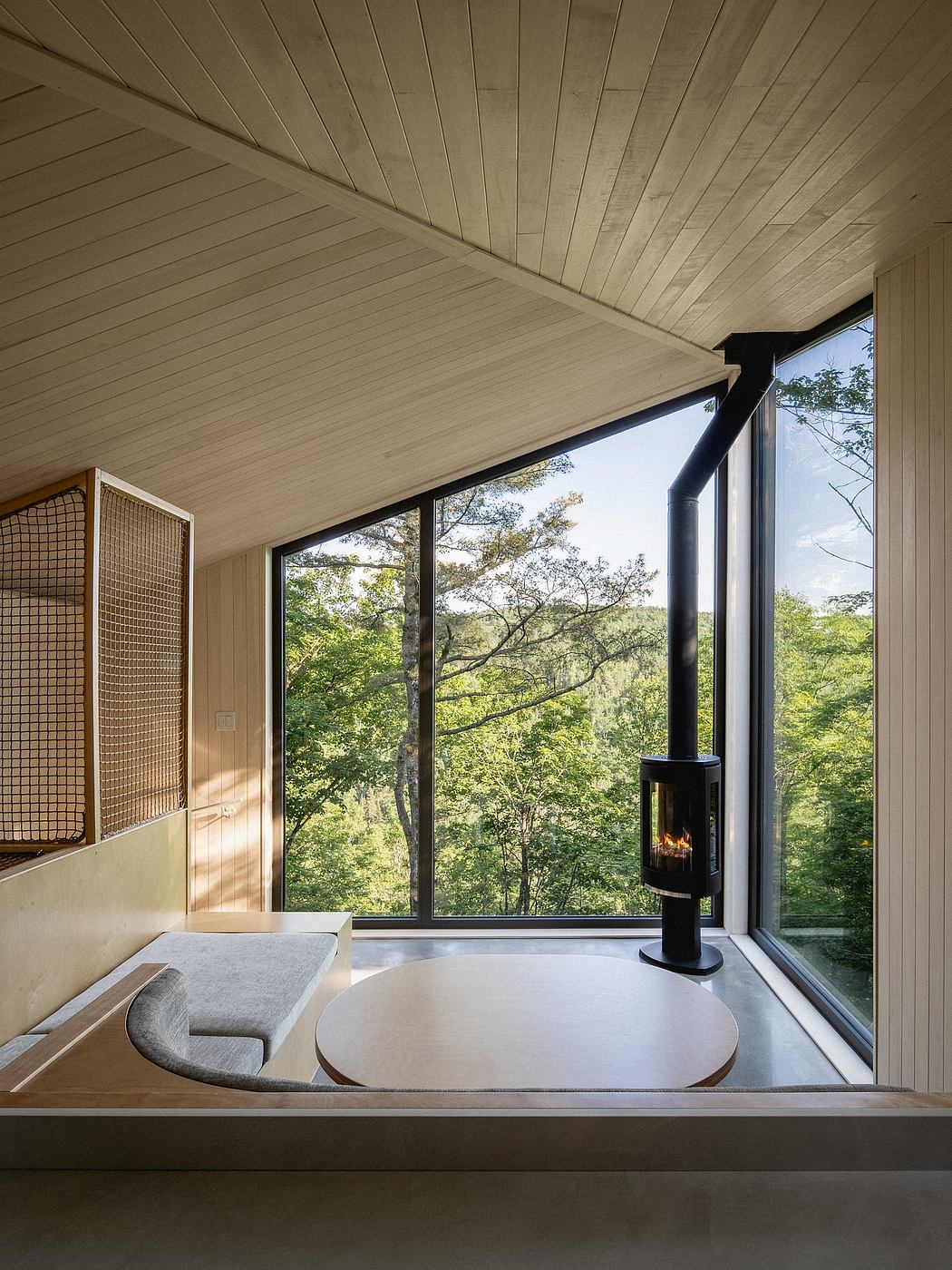 A cozy interior with a wood-paneled ceiling, large windows, and a modern fireplace overlooking the lush trees outside.