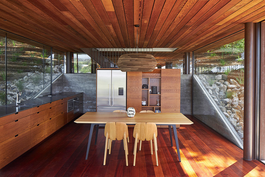 Warm-toned wood ceiling and walls, minimalist kitchen and dining area, glass walls.