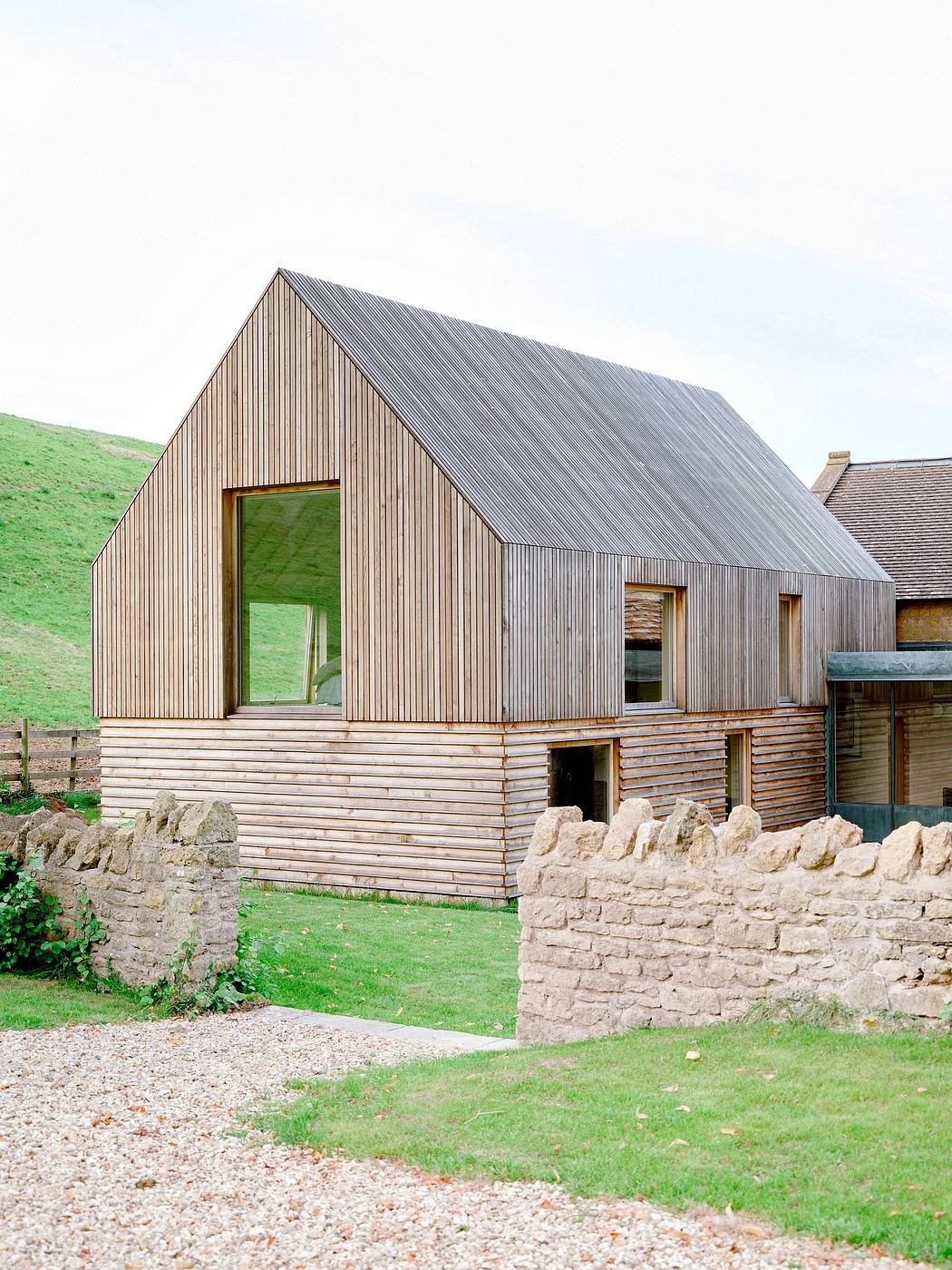 A modern wooden cabin with a slanted roof, surrounded by a stone wall and lush greenery.