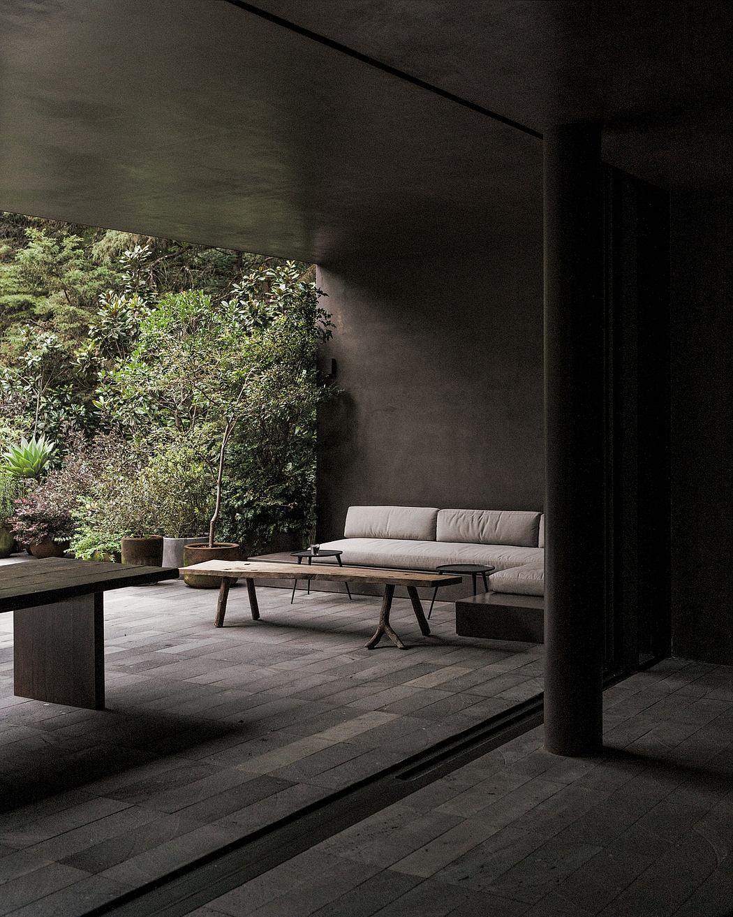 A dark, modern outdoor seating area with a couch, coffee tables, and lush vegetation.