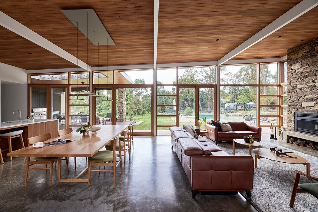 A modern, open-plan living space with wood-paneled ceilings, floor-to-ceiling windows, and a stone fireplace.