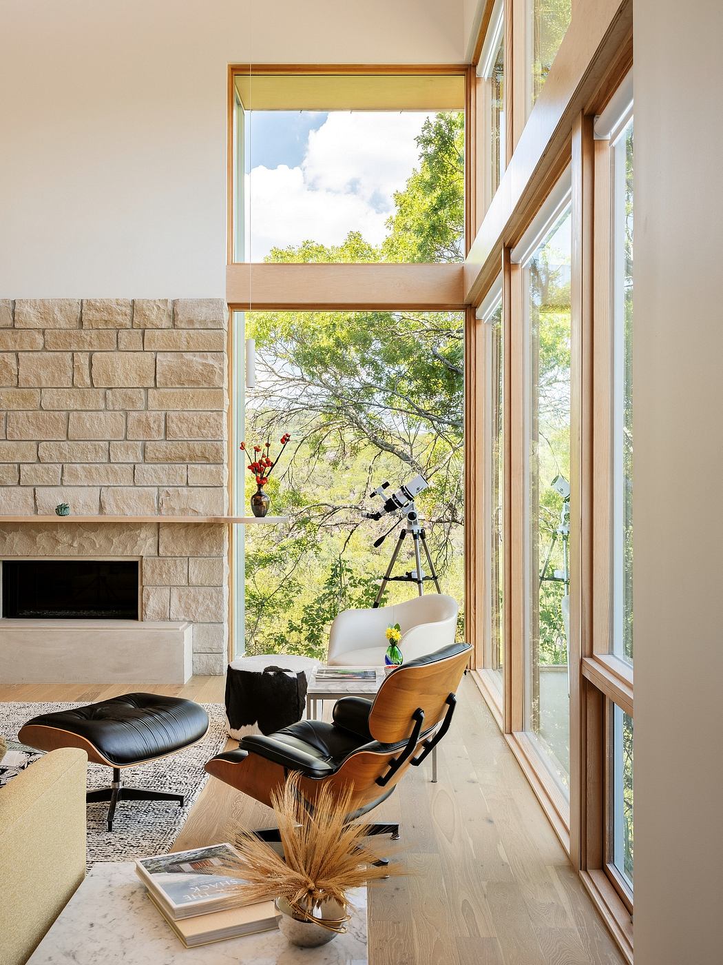 Spacious room with large windows, Eames chair, and telescope with a wooded backdrop.