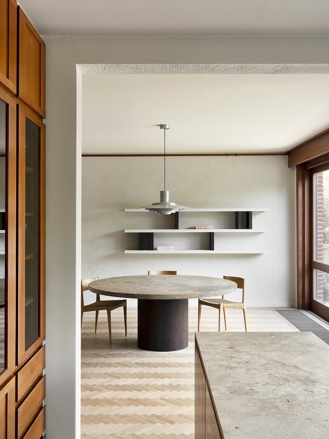 A minimalist dining room with a round wooden table, modern lighting, and built-in shelving.