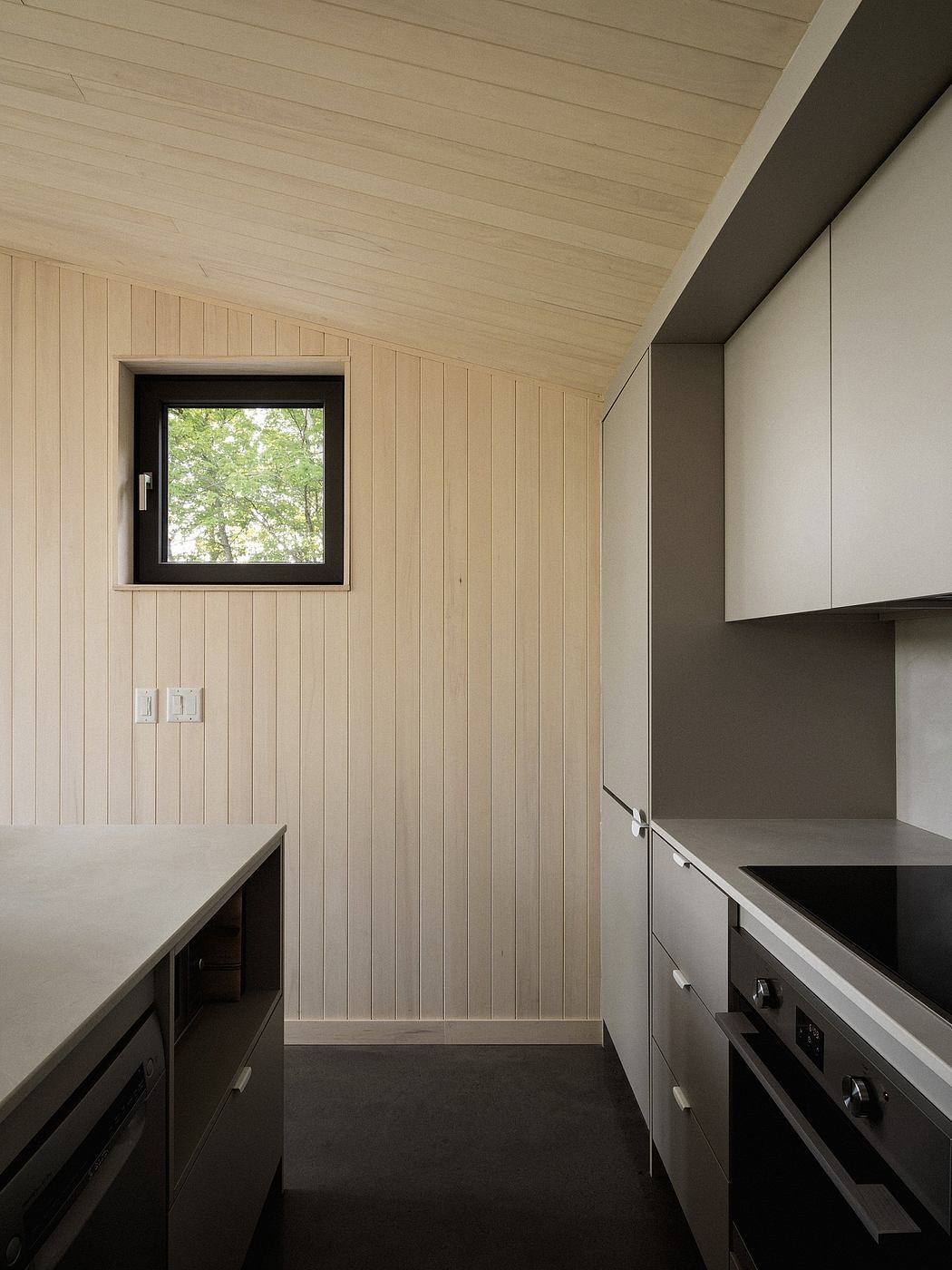 A cozy kitchen interior with wooden paneled walls, minimalist cabinetry, and a window overlooking greenery.