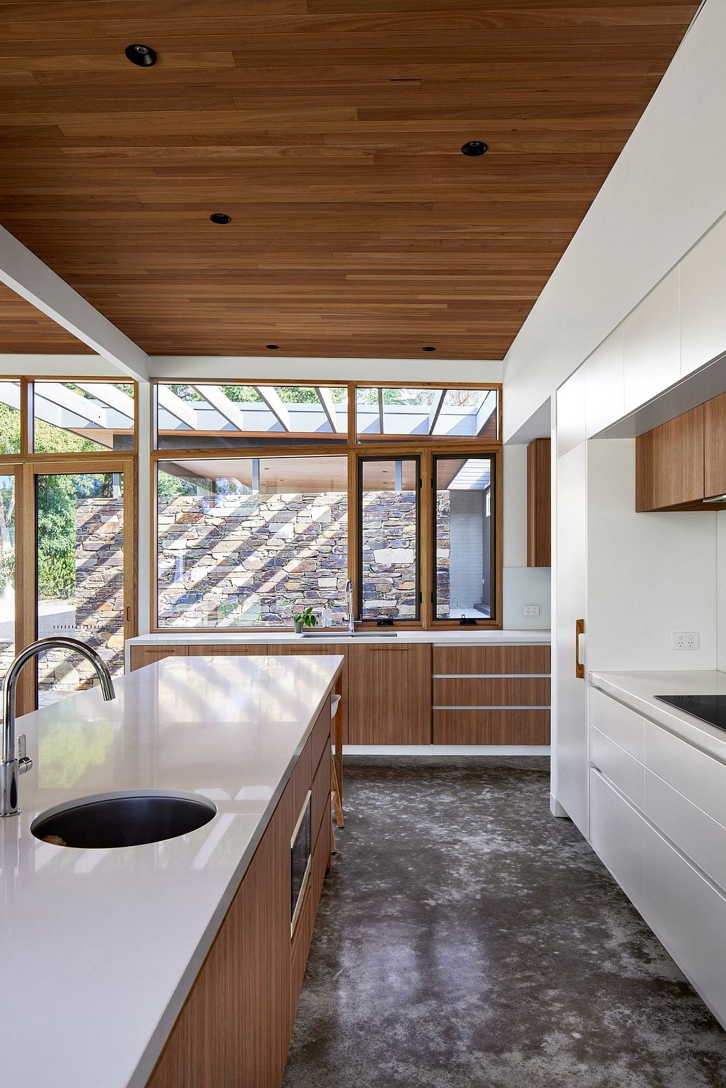 A modern kitchen with wooden ceiling, stone walls, and a concrete floor. Sleek cabinetry and large windows provide natural lighting.