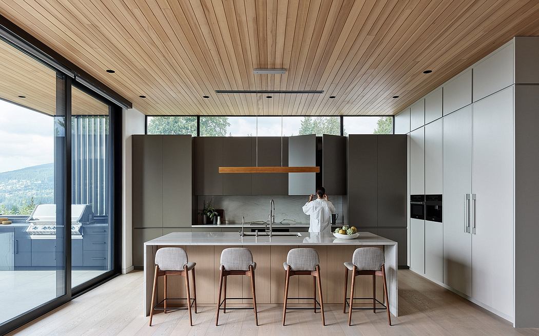 A modern, open-concept kitchen with sleek, dark cabinetry, wooden ceilings, and large windows offering scenic views.