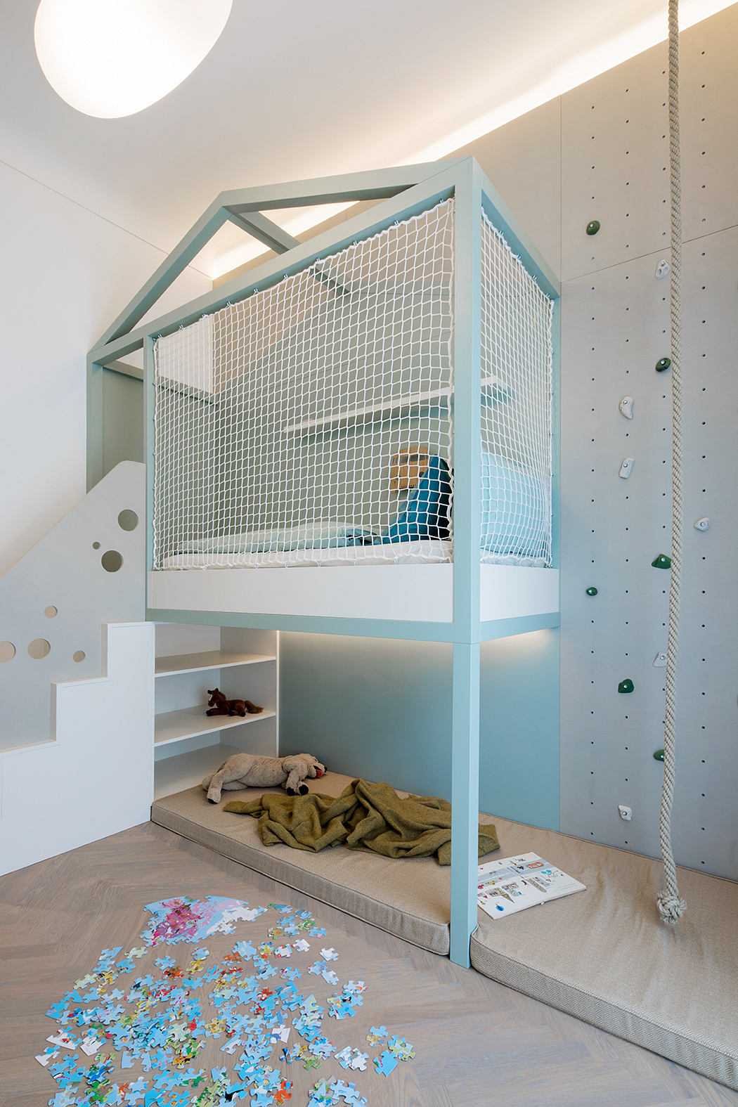 A modern children's room with a bunk bed encased in a mesh frame, climbing wall, and scattered puzzle pieces.