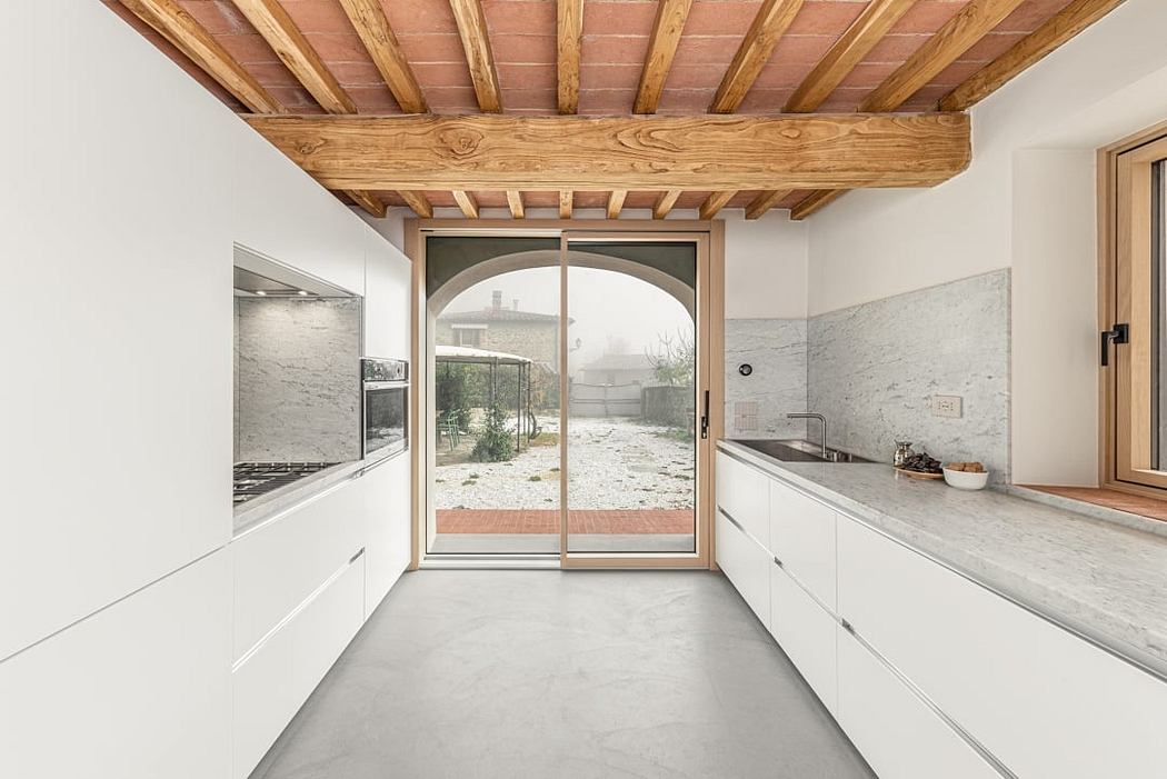 A modern kitchen with a rustic wood ceiling, white cabinets, and a large arched window overlooking an outdoor yard.