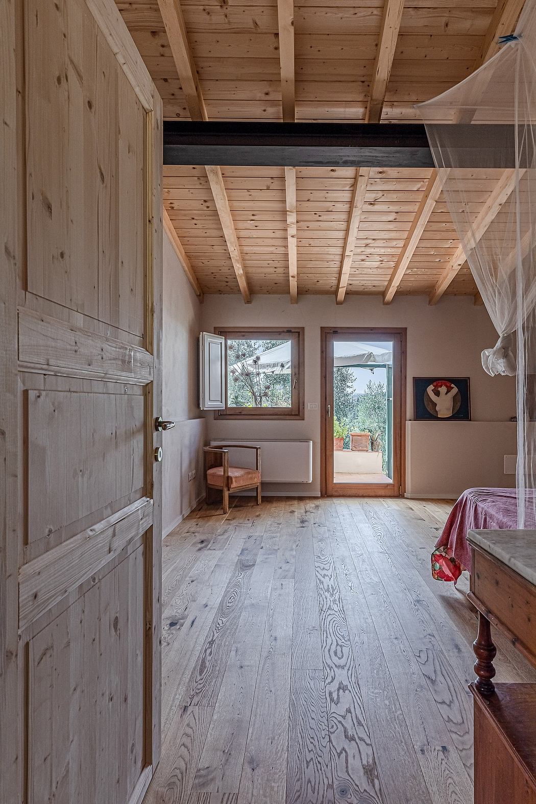 A cozy rustic bedroom with wooden beams, paneled walls, and a view to the outdoors.