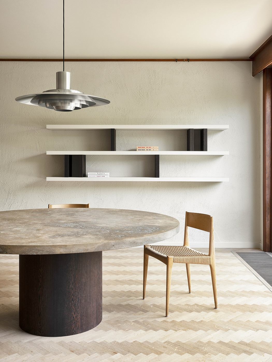 A modern dining space with a round stone table, woven wood chair, and minimalist shelving.